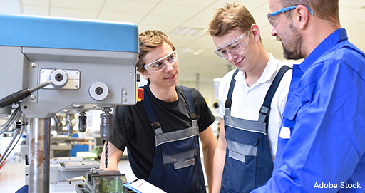 Zwei Lehrlinge und ein Lehrer stehen gemeinsam an einer Ständerbohrmaschine. Sie tragen Arbeitskleidung und Schutzbrillen.