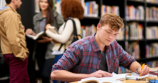 Junger Person sitzt lernend an einem Tisch in einer Bibliothek. Im Hintergrund unterhalten sich stehend drei Personen.
